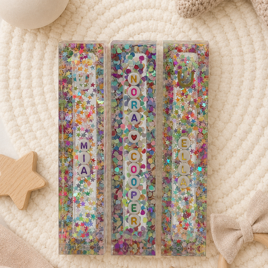 Three personalized glittery resin name cards with colourful confetti and letter accents, arranged on a cream woven mat with soft baby-themed props.