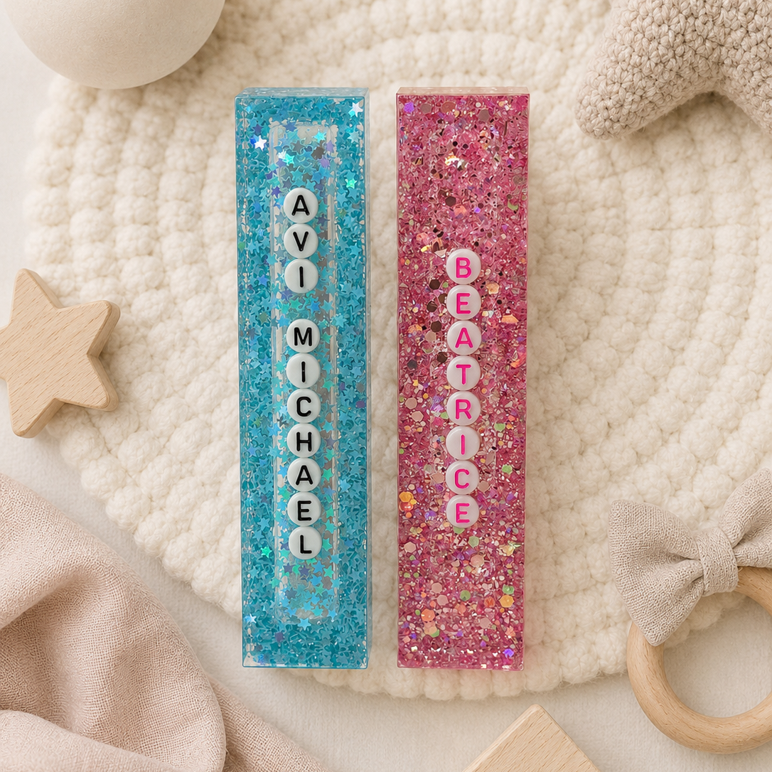 Two personalized glitter name bars for children, one in blue with the name 'AVI MICHAEL' and the other in pink with 'BEATRICE', styled on a cream knit blanket with baby toys.

