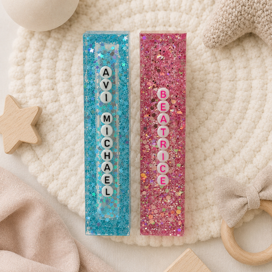 Two personalized glitter name bars for children, one in blue with the name 'AVI MICHAEL' and the other in pink with 'BEATRICE', styled on a cream knit blanket with baby toys.

